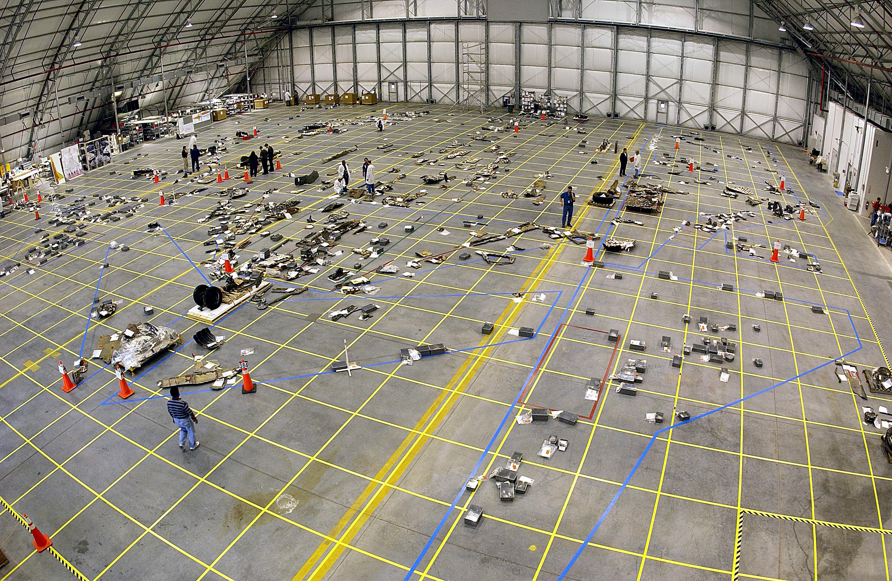 Umiestňovanie trosiek raketoplánu na vyznačené pozície na veľkom nákrese na podlahe hangáru. Na tomto obrázku je to umiestňovanie trosiek Columbie, ktorá havarovala vo februári 2003. Zdroj.