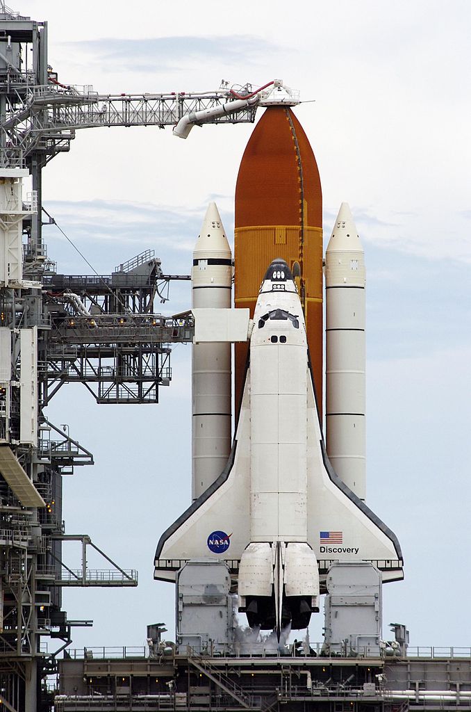 Discovery pred štartom z čelného pohľadu. Vlastný raketoplán (orbiter) je pripevnený k oranžovej nádrži ET, ktorá ho prevyšuje. Biele motory po bokoch pripomínajúce ceruzky sú motory SRB. Zdroj.